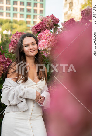 Young woman in a light dress stands among pink hydrangea flowers in an urban garden, soft daylight, pastel tones, calm and warm mood Young woman in a light dress stands among pink hydrangea flowers in an urban garden, soft daylight, pastel tones, calm and warm mood 134394386