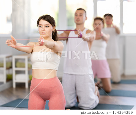 Concentrated sporty young girl exercising with group, kneeling on mat and performing exercise with pilates resistance bands in fitness studio 134394399
