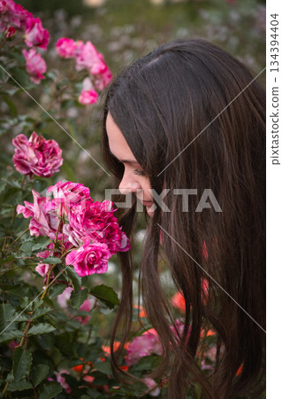 Young woman smells pink roses in a garden, close-up portrait, soft daylight, rich green and pink tones, gentle and contemplative mood 134394404