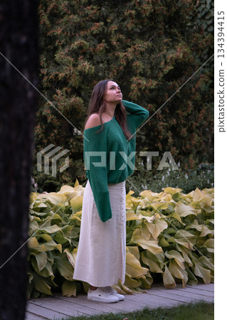 A woman in a green sweater and light skirt stands among lush garden plants, soft greenery, and muted natural tones, conveying calm and introspection 134394415