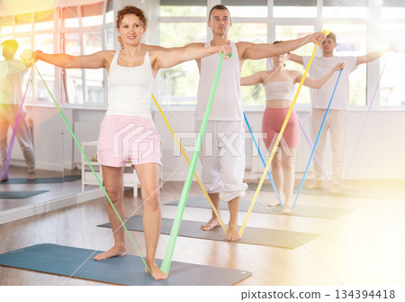 Positive woman with sporty women and men engaged in group class standing and perform exercise with resistance bands in pilates studio Positive woman with sporty women and men engaged in group class standing and perform exercise with resistance bands in pilates studio 134394418