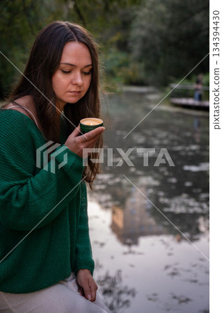 A woman holds a small lit candle in her hands, soft flame reflecting on her face against a blurred water and greenery background 134394430