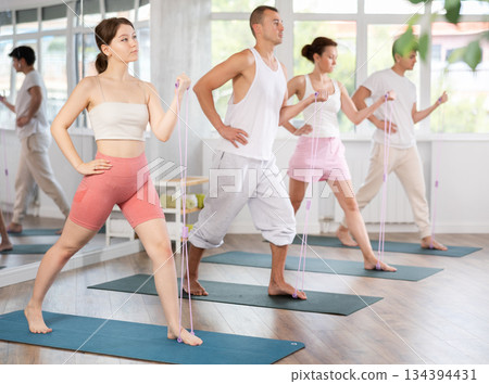 Sports focused young woman training with resistance band with group, strong biceps or abs muscles in gym health center 134394431