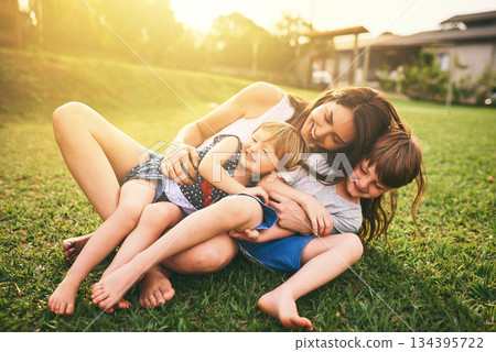 Mother, children and hug playing on grass for fun bonding in the sun outside their house in nature. Happy mom hugging kids on garden floor outdoors in playful, joy and happiness of family together 134395722