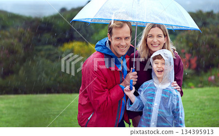 Family, happy and portrait in rain with umbrella in nature outdoor for fun, happiness and quality time. Man, woman and boy child with protection for water drops with freedom while playing in winter Family, happy and portrait in rain with umbrella in nature outdoor for fun, happiness and quality time. Man, woman and boy child with protection for water drops with freedom while playing in winter 134395913