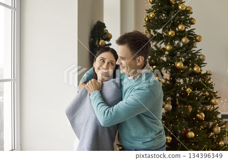 Happy young couple standing at home and hugging covered in cozy blanket on Christmas eve. 134396349