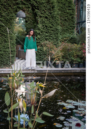 A young woman in a green sweater and white trousers stands by a small pond on a wooden deck, surrounded by dense garden greenery and calm reflections 134396419