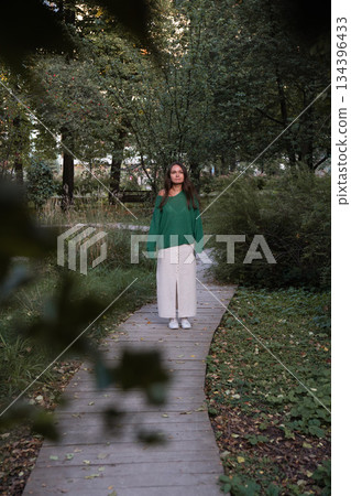 A woman in a green sweater and light skirt walks along a narrow wooden path surrounded by lush greenery, creating a calm and intimate nature scene A woman in a green sweater and light skirt walks along a narrow wooden path surrounded by lush greenery, creating a calm and intimate nature scene 134396433