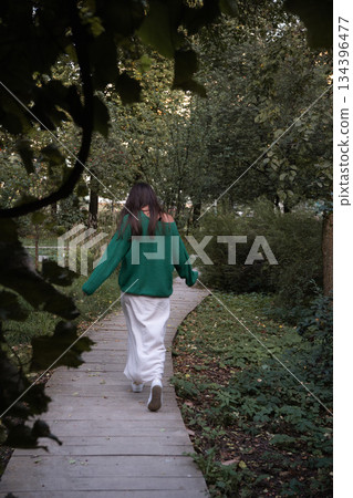 Woman in a green sweater and light skirt walks along a wooden path surrounded by dense greenery, captured from behind with a sense of motion Woman in a green sweater and light skirt walks along a wooden path surrounded by dense greenery, captured from behind with a sense of motion 134396477