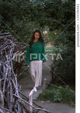 A woman in a green sweater and light skirt walks along a narrow wooden path surrounded by lush greenery, creating a calm and intimate nature scene 134396481