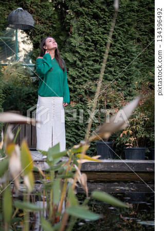 A young woman in a green sweater and white trousers stands by a small pond on a wooden deck, surrounded by dense garden greenery and calm reflections A young woman in a green sweater and white trousers stands by a small pond on a wooden deck, surrounded by dense garden greenery and calm reflections 134396492