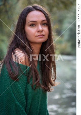 A calm portrait of a woman in a green sweater standing by a quiet pond, with soft natural light, blurred greenery 134396497