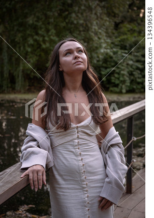 Young woman in a light dress and cardigan stands on a wooden bridge by a pond, looking upward, soft daylight, muted green tones, reflective mood 134396548