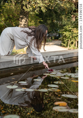 Woman reaches toward floating leaves on calm pond water from a wooden edge, soft daylight, warm reflections, muted green and brown tones 134396558