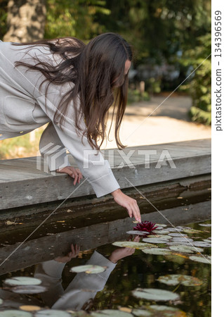 Woman reaches toward floating leaves on calm pond water from a wooden edge, soft daylight, warm reflections, muted green and brown tones Woman reaches toward floating leaves on calm pond water from a wooden edge, soft daylight, warm reflections, muted green and brown tones 134396569