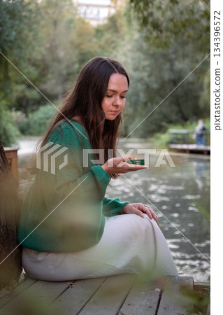 Young woman in a green sweater holds a small lit candle while sitting by a calm pond, soft daylight, muted green tones, quiet and contemplative mood 134396572