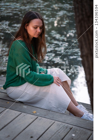 Young woman sits on a wooden deck by the water, looking down, soft natural light, muted green and gray tones, quiet and introspective mood 134396594