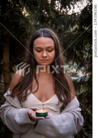 Young woman in a dress and cardigan holds a small lit candle with eyes closed among evergreen trees, soft daylight, deep green tones, centered mood 134396596