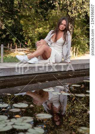 Young woman in a light dress sits on a wooden edge by a calm pond with reflections and floating leaves, soft daylight, warm natural tones, serene mood 134396607