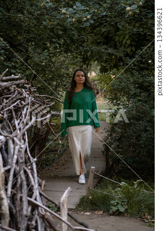 A woman in a green sweater and light skirt walks along a narrow wooden path surrounded by lush greenery, creating a calm and intimate nature scene 134396621