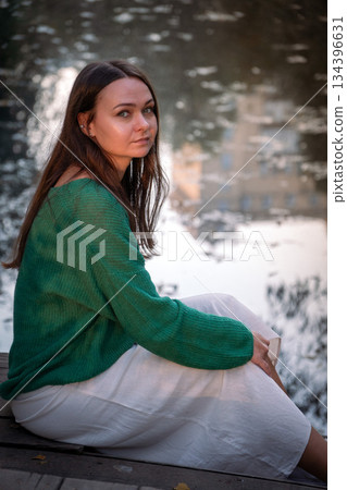 Young woman sits on a wooden deck by the water, looking down, soft natural light, muted green and gray tones, quiet and introspective mood Young woman sits on a wooden deck by the water, looking down, soft natural light, muted green and gray tones, quiet and introspective mood 134396631