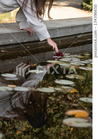 Woman reaches toward floating leaves on calm pond water from a wooden edge, soft daylight, warm reflections, muted green and brown tones Woman reaches toward floating leaves on calm pond water from a wooden edge, soft daylight, warm reflections, muted green and brown tones 134396650