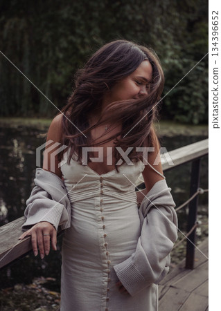 Young woman in a dress and cardigan stands on a wooden bridge by a pond, wind moving her hair, soft daylight, muted green tones, spontaneous mood Young woman in a dress and cardigan stands on a wooden bridge by a pond, wind moving her hair, soft daylight, muted green tones, spontaneous mood 134396652