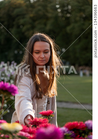 Young woman in a light dress reaches toward pink roses in a park garden, soft evening light, warm highlights, green background, calm and inviting mood Young woman in a light dress reaches toward pink roses in a park garden, soft evening light, warm highlights, green background, calm and inviting mood 134396663
