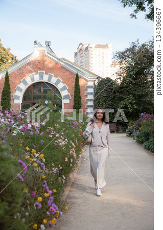 A woman walks along a garden path beside blooming flowers, historic pavilion and city buildings in the background, daylight 134396667