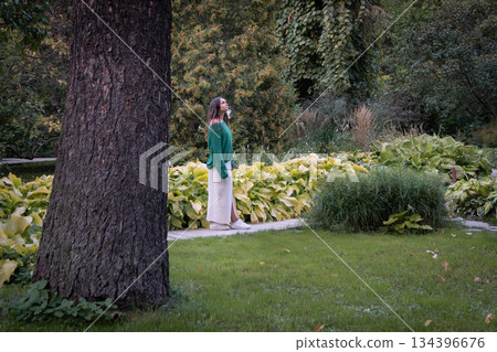 A woman in a green sweater and light skirt stands in a lush garden, surrounded by tall ivy-covered trees, leafy plants, and soft natural light 134396676