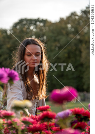Young woman in a light dress reaches toward pink roses in a park garden, soft evening light, warm highlights, green background, calm and inviting mood Young woman in a light dress reaches toward pink roses in a park garden, soft evening light, warm highlights, green background, calm and inviting mood 134396682