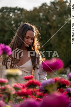 Young woman in a light dress reaches toward pink roses in a park garden, soft evening light, warm highlights, green background, calm and inviting mood Young woman in a light dress reaches toward pink roses in a park garden, soft evening light, warm highlights, green background, calm and inviting mood 134396687