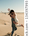 Man lifting woman joyfully on a sandy beach, both smiling in a playful and loving moment under clear skies. 134397993