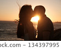 Couple sharing a romantic kiss at sunset by the sea, silhouetted against the golden light of the setting sun. 134397995