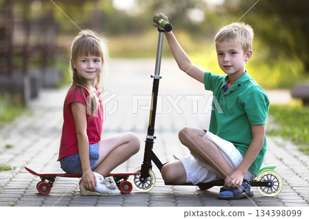 Two cute happy funny smiling young children, brother and sister, posing for camera, handsome boy with scooter and pretty long-haired girl sitting on skateboard on sunny colorful blurred background. 134398099