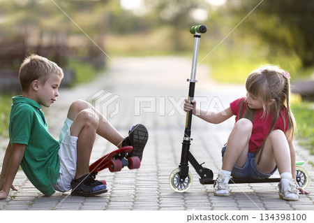 Two cute happy funny laughing young children, brother and sister, having fun on sunny pavement, handsome boy with skateboard and pretty long-haired girl fallen from scooter on blurred background. 134398100