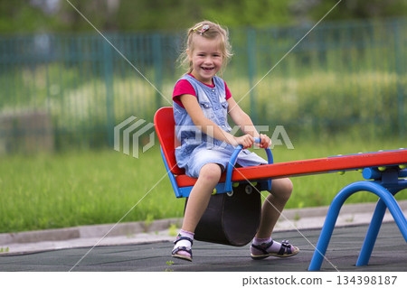 Small cute young blond child girl sits on see- saw swing on warm sunny day on bright green blurred background. Joys and sorrows of childhood, outdoor activity concept. 134398187