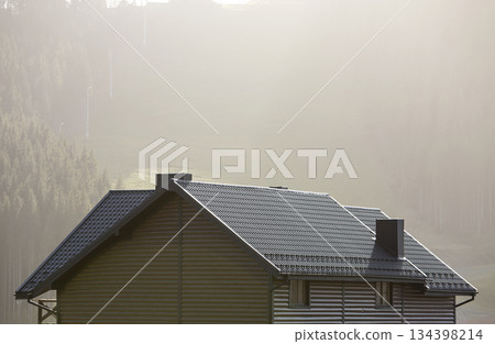 Roof of cottage with siding walls, brown shingle roof and high chimneys in ecological area on foggy landscape copy space background on sunny summer day. Well maintained property, real estate concept. 134398214