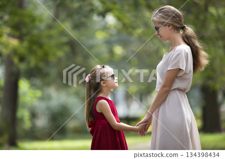 Profile of young blond long-haired attractive smiling woman and small child girl in sunglasses and fashionable dresses holding hands looking on each other on sunny summer green bokeh background. 134398434