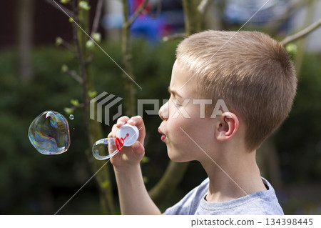 Profile of cute handsome small blond child boy with funny serious expression blowing colorful transparent soap bubbles outdoors on blurred green summer background. Joy of careless childhood concept. Profile of cute handsome small blond child boy with funny serious expression blowing colorful transparent soap bubbles outdoors on blurred green summer background. Joy of careless childhood concept. 134398445