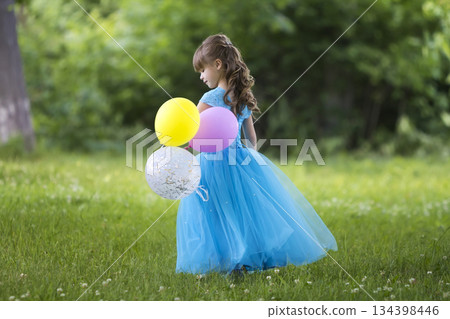 Profile full-length portrait of pretty little blond long-haired girl in long blue dress with colorful balloons standing in blooming field on blurred green trees background. Happy childhood concept. 134398446