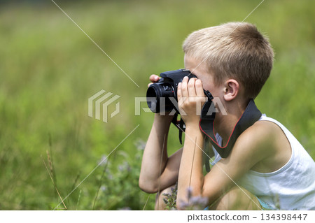 Profile close-up portrait of young blond cute handsome child boy with camera taking pictures outdoors on bright sunny spring or summer day on blurred light green grassy copy space background. 134398447
