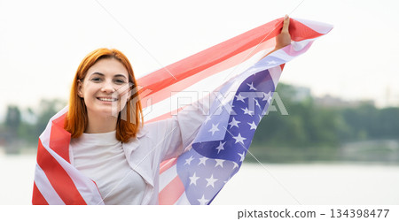 Portrait of happy smiling red haired girl with USA national flag on her shoulders. Positive young woman celebrating United States independence day. Portrait of happy smiling red haired girl with USA national flag on her shoulders. Positive young woman celebrating United States independence day. 134398477