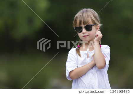 Portrait of cute pretty funny young girl with blond braids in white dress and dark sunglasses. 134398485