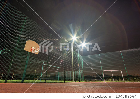 Outdoors mini football and basketball court with ball gate and basket surrounded with high protective fence brightly illuminated with spotlight lamps at night. 134398564