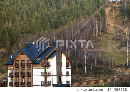 New modern comfortable four-story hotel building with attached premises, attic rooms and high chimneys in ecological rural area on spruce dense forest. Recreation and tourism concept. New modern comfortable four-story hotel building with attached premises, attic rooms and high chimneys in ecological rural area on spruce dense forest. Recreation and tourism concept. 134398588