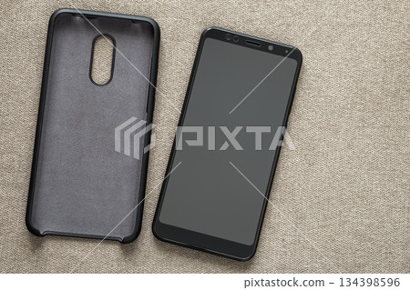 New black modern cellphone and plastic protective case isolated on light cloth copy space background. Modern technology, communication, gadget design and accessories concept. New black modern cellphone and plastic protective case isolated on light cloth copy space background. Modern technology, communication, gadget design and accessories concept. 134398596
