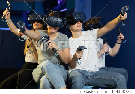 group of four friends wearing virtual reality glasses and with joysticks sit and play dynamic game group of four friends wearing virtual reality glasses and with joysticks sit and play dynamic game 134398785