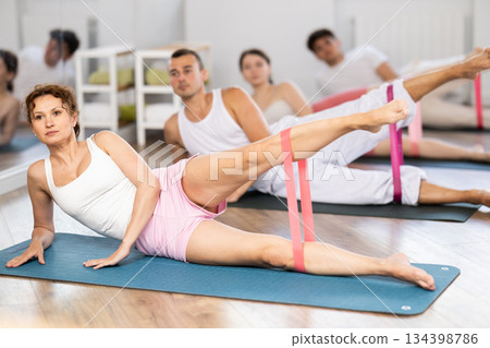 Professional active woman exercising with resistance bands lying on side and stretching legs during group pilates class at gym Professional active woman exercising with resistance bands lying on side and stretching legs during group pilates class at gym 134398786