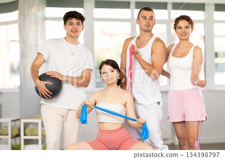 Young men and women posing together in training room holding resistance bands and pilates ball Young men and women posing together in training room holding resistance bands and pilates ball 134398797